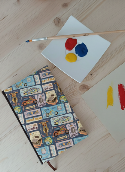 Carnet à la couverture imprimé de dessins de voitures, motos et panneaux de la route 66 à côté d'une palette de peinture et un pinceau.
