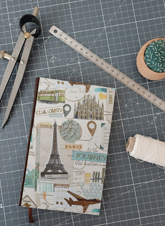 Carnet à la couverture imprimée de dessins de monuments et tampons de voyage, entouré d'un compas, d'un réglet d'aiguilles et de fil.