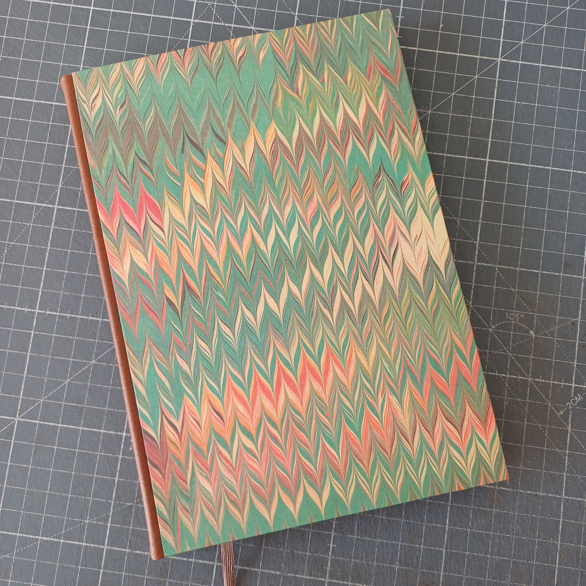 Carnet artisanal avec une couverture imprimée d'un marbré en forme de chevrons orangés et verts, avec un dos en similicuir marron et un signet marron.