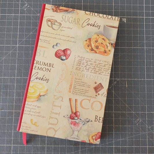 Carnet artisanal à la couverture imprimée de motifs de pâtisserie, de fruits et de café avec des écritures évoquant des recettes, et un dos en similicuir rouge.