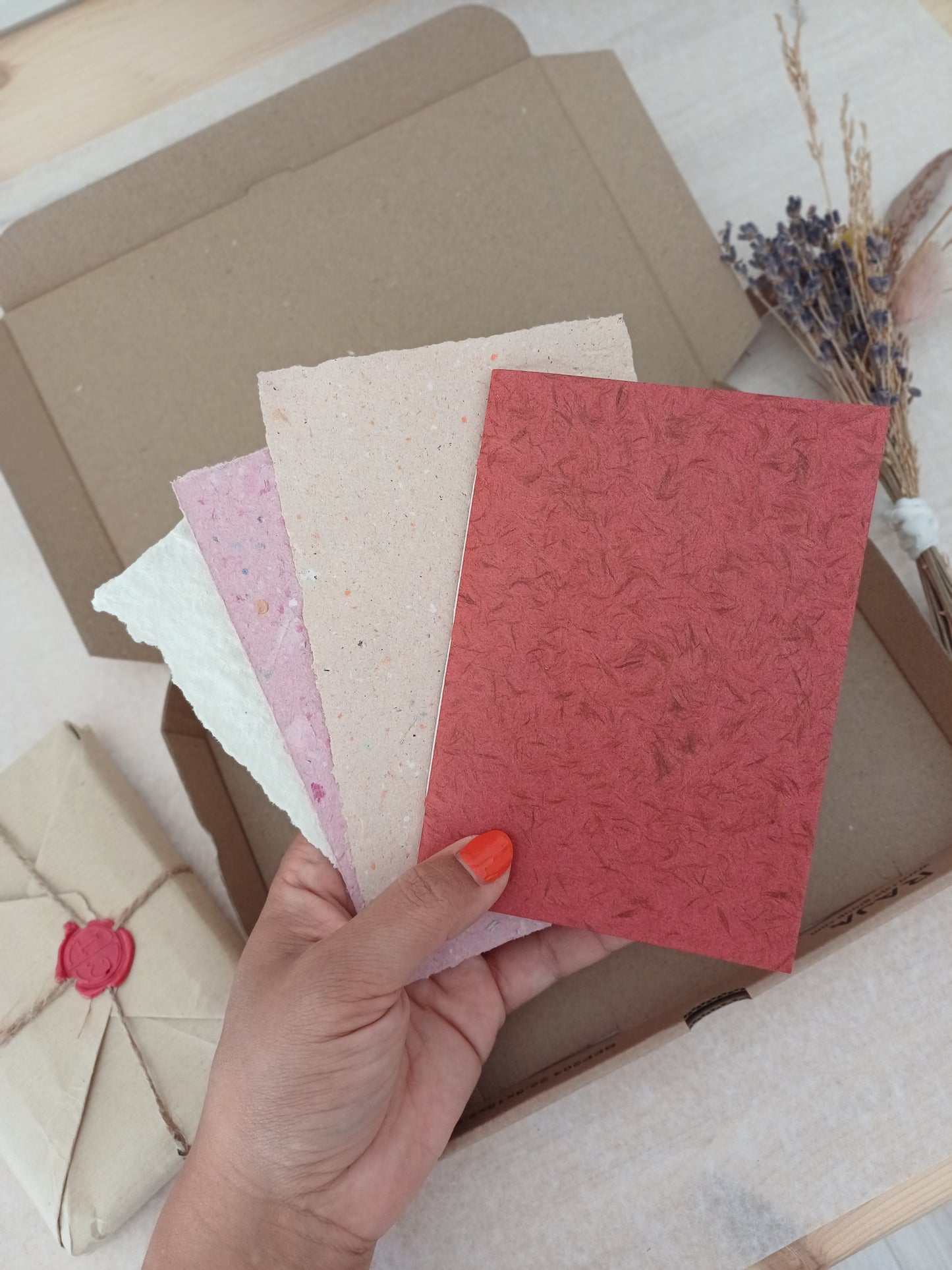 Main tenant un petit carnet rouge avec des feuilles de papier recyclé devant une boite en carton, des fleurs séchées et un paquet cadeau au style vintage.