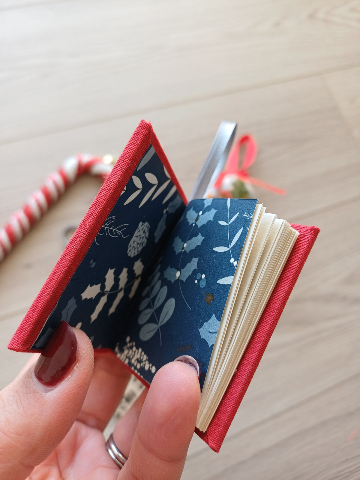 Un petit carnet rouge tenue par une main et entrouvert où l'on voit des pages de garde aux feuillages de Noël.