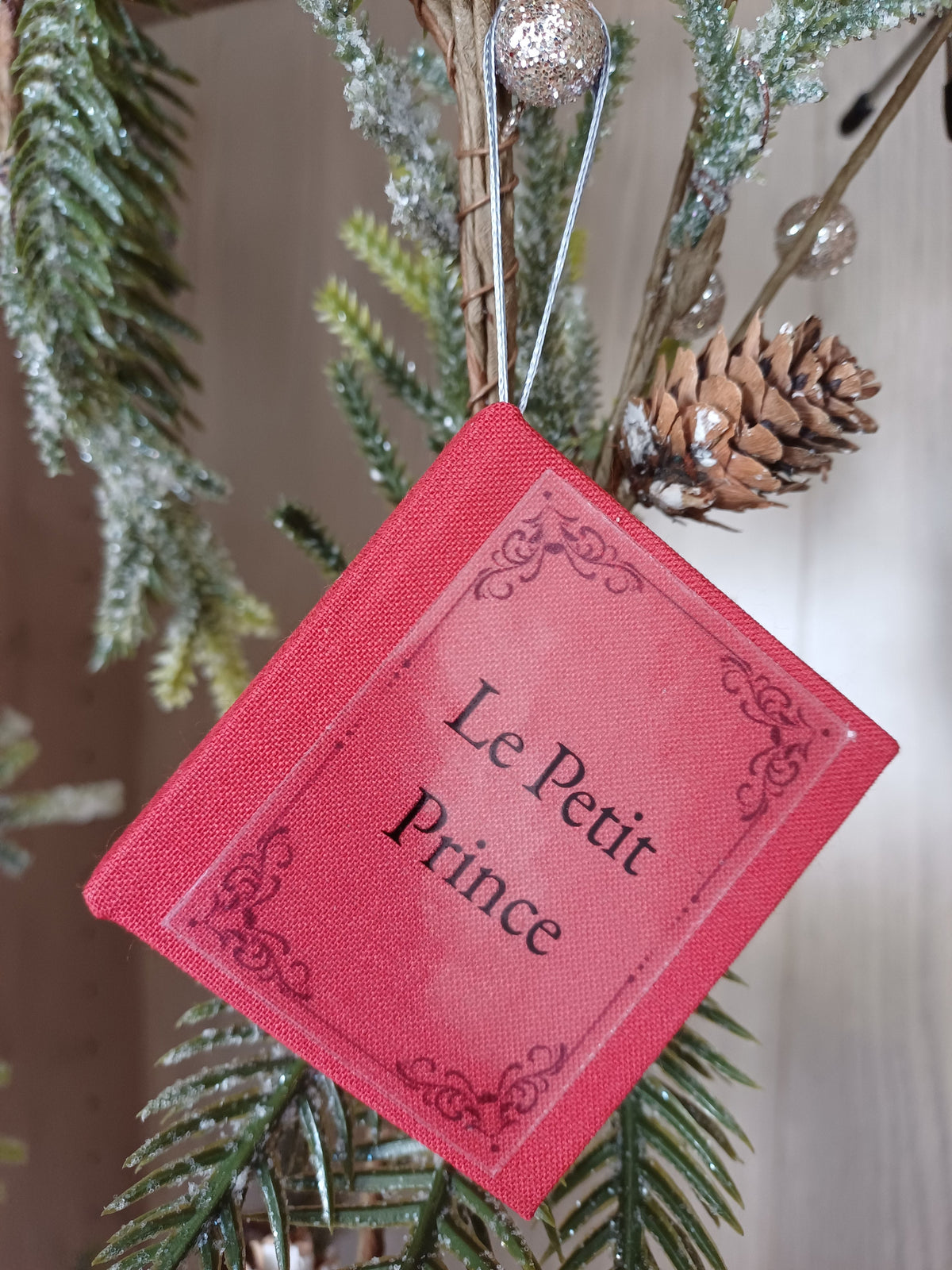 Un petit carnet rouge en forme de livre avec le tire "Le Petit Prince" accroché à une branche de sapin.