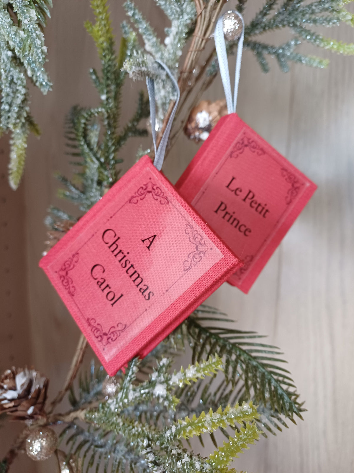 Deux petits carnets rouge en forme de livre avec les tire "A Christmas Carol" et "Le Petit Prince" accrochés à une branche de sapin de Noël.