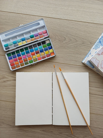 Carnet artisanal pour aquarelle ouvert avec deux pinceaux posés dessus et une palette de peinture.