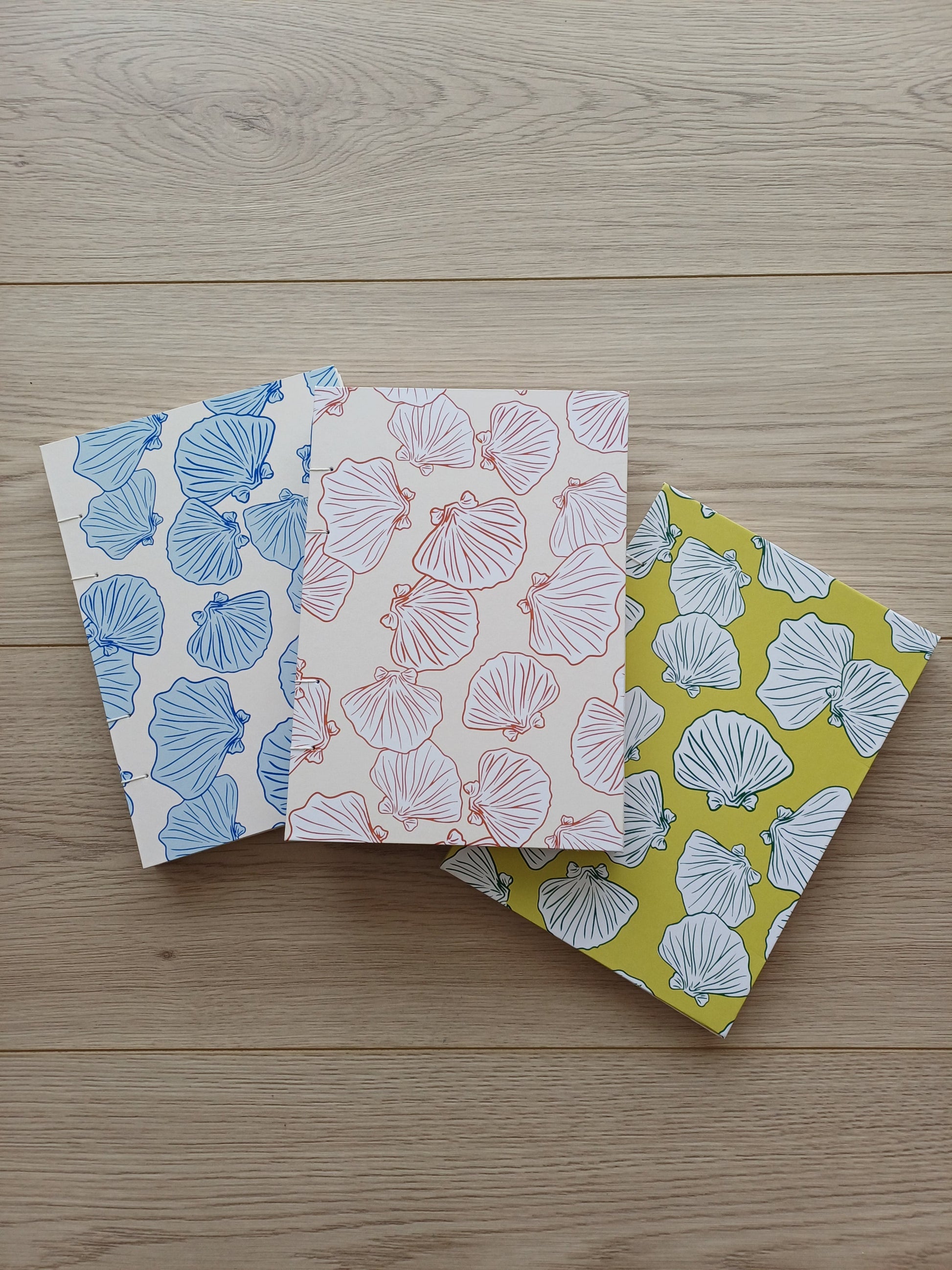 Couverture de trois carnets artisanaux pour aquarelle avec des motifs de coquillages sur des fonds vert, terracotta et bleu.