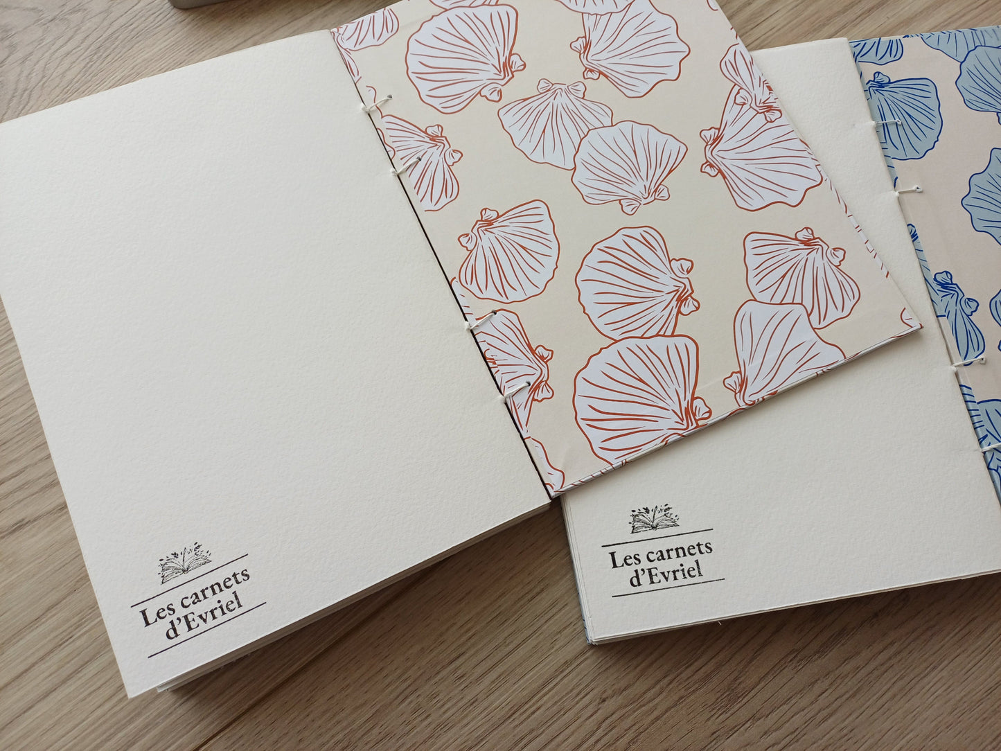 Carnets artisanaux pour aquarelle ouverts sur la dernière page avec un tampon "Les carnets d'Evriel" et la page de garde aux motifs coquillages.