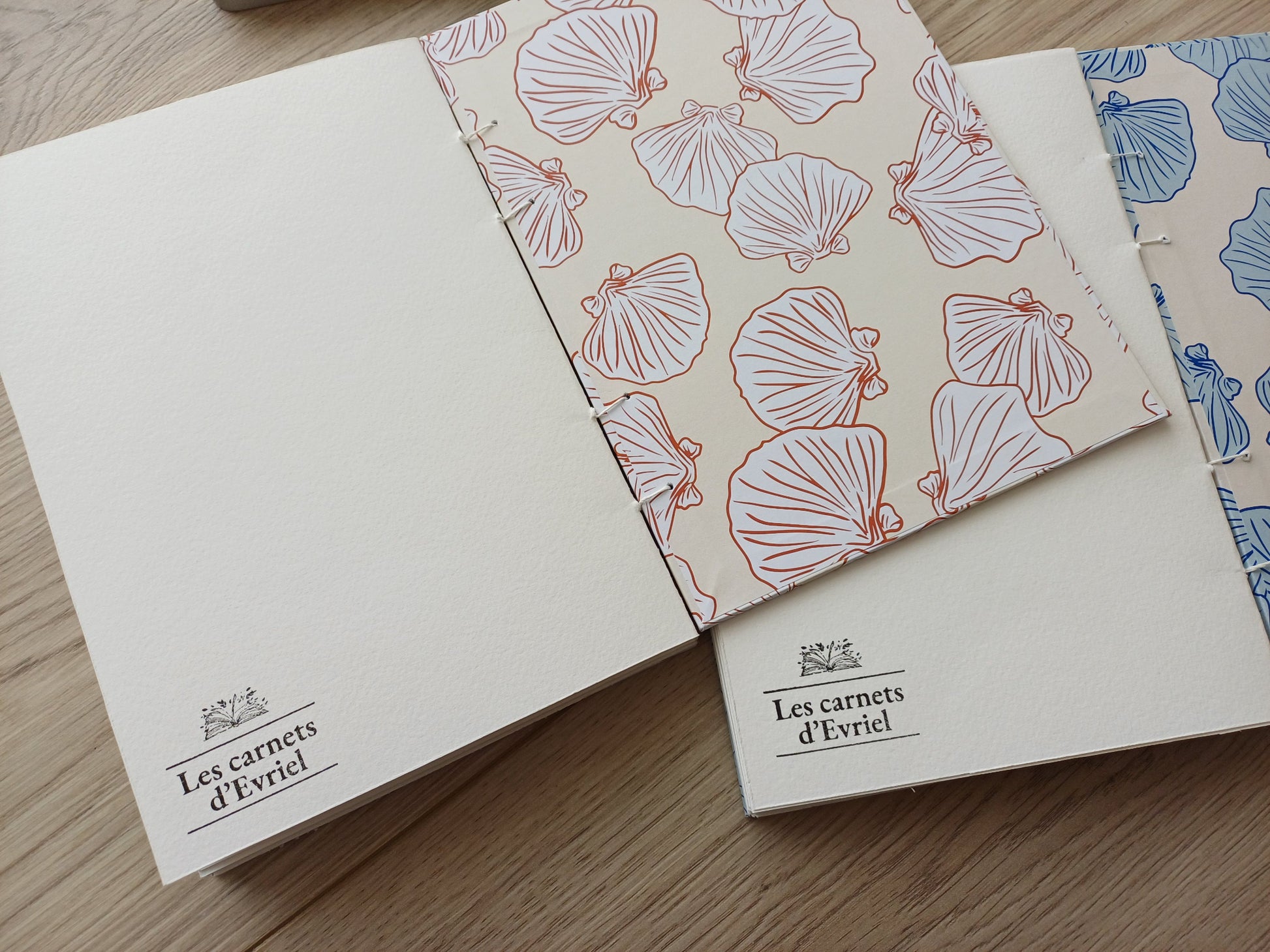 Carnets artisanaux pour aquarelle ouverts sur la dernière page avec un tampon "Les carnets d'Evriel" et la page de garde aux motifs coquillages.