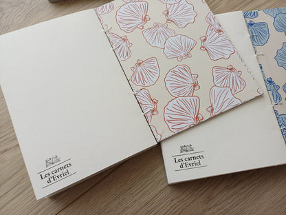 Carnets artisanaux pour aquarelle ouverts sur la dernière page avec un tampon "Les carnets d'Evriel" et la page de garde aux motifs coquillages.